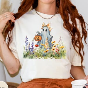 May include: Ivory-colored t-shirt featuring a watercolor illustration of a ghost holding a jack-o'-lantern in a field of wildflowers. The ghost wears a bow and is surrounded by purple, yellow, and red flowers. The shirt is paired with orange shorts.