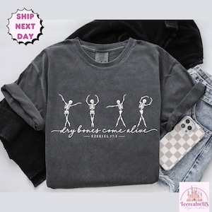May include: A charcoal gray t-shirt featuring a white graphic of four dancing skeletons. Below the skeletons, the text reads "dry bones come alive" in a cursive script, followed by the printed reference "EZEKIEL 37:5" in a clean font.