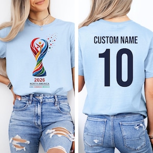 Op de afbeelding: Lichtblauw t-shirt met een kleurrijk ontwerp en de tekst "2026 NORTH AMERICA USA CANADA MEXICO" op de voorkant. De achterkant van het shirt toont "CUSTOM NAME" boven het nummer "10" in marineblauw.