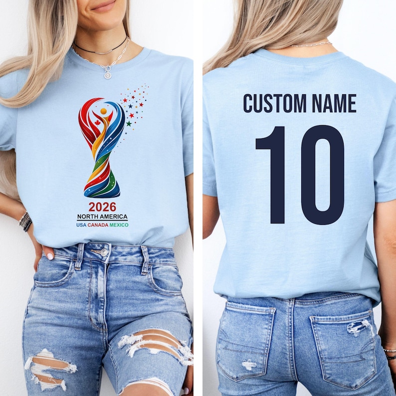 Op de afbeelding: Lichtblauw t-shirt met een kleurrijk ontwerp en de tekst "2026 NORTH AMERICA USA CANADA MEXICO" op de voorkant. De achterkant van het shirt toont "CUSTOM NAME" boven het nummer "10" in marineblauw.
