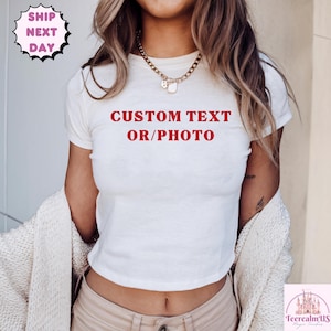 May include: A white short-sleeved t-shirt featuring the red text CUSTOM TEXT OR/PHOTO printed on the chest. The shirt has a crew neckline and is worn with a gold-toned chain necklace and a cream-colored textured cardigan draped over the shoulders.
