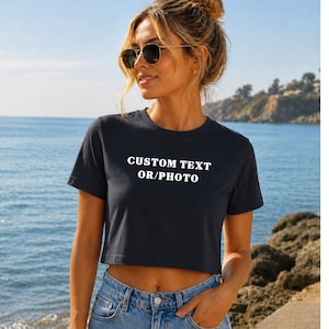 A woman wearing a black crop top with the text "CUSTOM TEXT OR PHOTO" stands near a rocky beach with the ocean in the background.