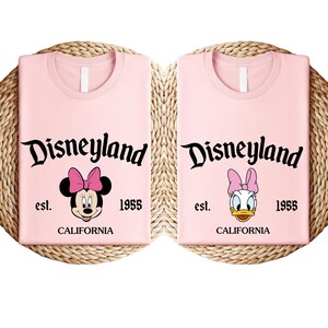 May include: Two pale pink t-shirts with the word "Disneyland" in black script. One shirt features Minnie Mouse, the other Daisy Duck. Both shirts have "est. 1955 CALIFORNIA" printed below the characters.