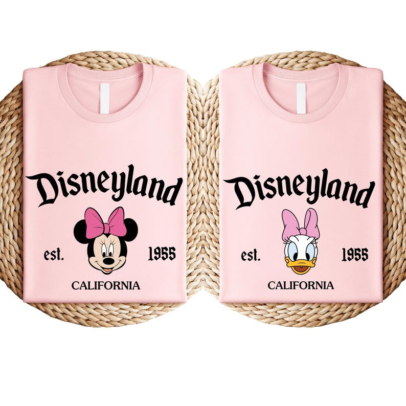 May include: Two pale pink t-shirts with the word "Disneyland" in black script. One shirt features Minnie Mouse, the other Daisy Duck. Both shirts have "est. 1955 CALIFORNIA" printed below the characters.