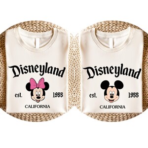 May include: Two cream-coloured t-shirts with the Disneyland logo and the text "est. 1955 CALIFORNIA". One shirt features Minnie Mouse with a pink bow, the other features Mickey Mouse. The shirts are folded on a woven surface.