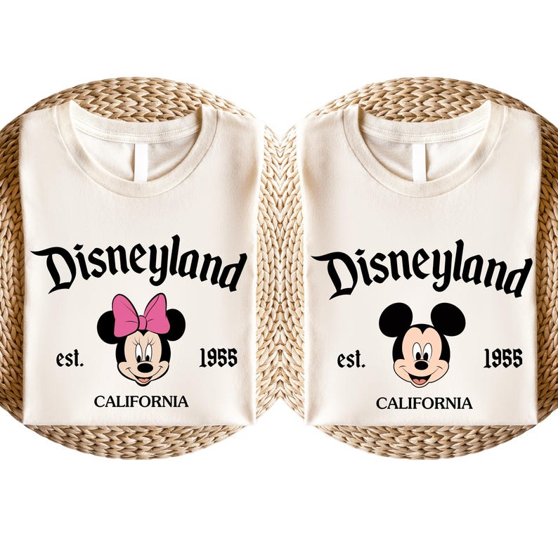May include: Two cream-coloured t-shirts with the Disneyland logo and the text "est. 1955 CALIFORNIA". One shirt features Minnie Mouse with a pink bow, the other features Mickey Mouse. The shirts are folded on a woven surface.