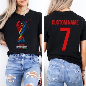 Op de afbeelding: Zwart T-shirt met het kleurrijke logo van het Wereldkampioenschap voetbal 2026 in Noord-Amerika. De achterkant van het shirt toont "CUSTOM NAME" en het nummer 7 in rood. Het shirt wordt gedragen met een blauwe jeans.