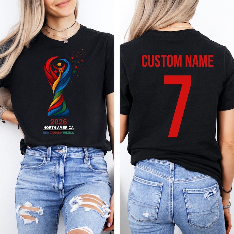 Op de afbeelding: Zwart T-shirt met het kleurrijke logo van het Wereldkampioenschap voetbal 2026 in Noord-Amerika. De achterkant van het shirt toont "CUSTOM NAME" en het nummer 7 in rood. Het shirt wordt gedragen met een blauwe jeans.