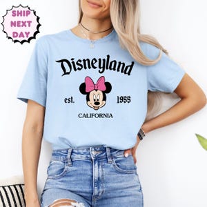 May include: Pale blue t-shirt featuring the word "Disneyland" in black script. Below the text is a Minnie Mouse graphic with a pink bow. The shirt also has the text "est. 1955 CALIFORNIA".
