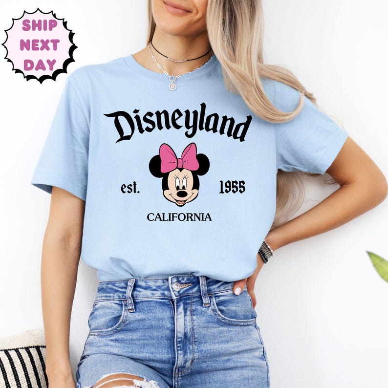 May include: Pale blue t-shirt featuring the word "Disneyland" in black script. Below the text is a Minnie Mouse graphic with a pink bow. The shirt also has the text "est. 1955 CALIFORNIA".