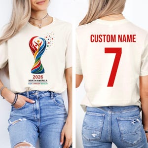 Op de afbeelding: Cr&egrave;mekleurig T-shirt met een kleurrijke afbeelding van een trofee en de tekst "2026 NORTH AMERICA USA CANADA MEXICO." De achterkant van het shirt toont "CUSTOM NAME" en het nummer "7" in rood.