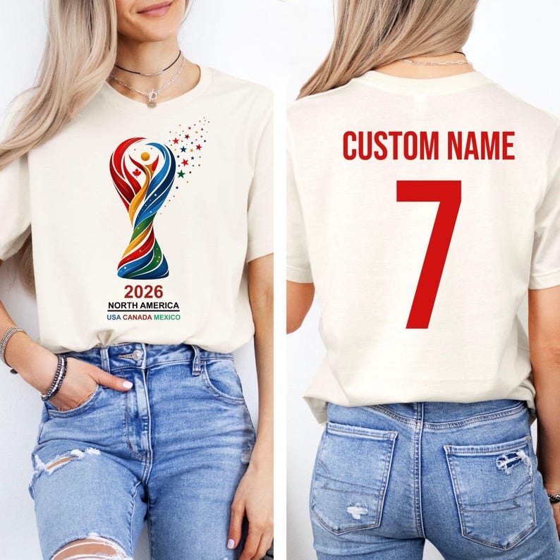 Op de afbeelding: Cr&egrave;mekleurig T-shirt met een kleurrijke afbeelding van een trofee en de tekst "2026 NORTH AMERICA USA CANADA MEXICO." De achterkant van het shirt toont "CUSTOM NAME" en het nummer "7" in rood.