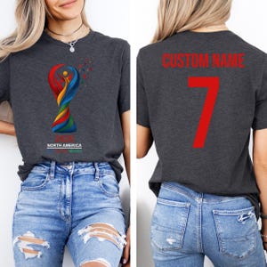Op de afbeelding: Donkergrijs t-shirt met een kleurrijke afbeelding van een trofee en de tekst "NORTH AMERICA MEXICO". De achterkant van het shirt heeft de tekst "CUSTOM NAME" en het nummer 7 in rood.