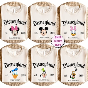 May include: Cream-coloured t-shirts featuring the Disneyland logo with various Disney character graphics. Each shirt has the text "est. 1955 CALIFORNIA". Characters include Minnie Mouse, Mickey Mouse, Daisy Duck, Donald Duck, Goofy, and Pluto.