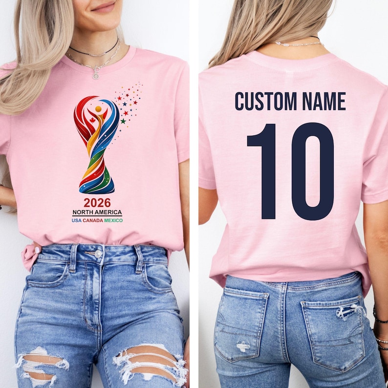 Op de afbeelding: Lichtroze T-shirt met een kleurrijk ontwerp van het logo van het WK 2026 in Noord-Amerika. De voorkant toont het logo en de tekst, terwijl de achterkant het nummer 10 en de tekst "CUSTOM NAME" in marineblauw heeft.