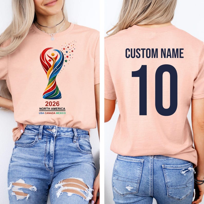 Op de afbeelding: Lichtperzikkleurig T-shirt met een kleurrijke trofee-afbeelding en de tekst "2026 NORTH AMERICA USA CANADA MEXICO". De achterkant van het shirt toont "CUSTOM NAME" boven het nummer "10" in marineblauw.