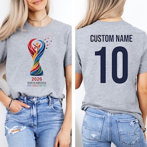 Op de afbeelding: Grijs t-shirt met een kleurrijke trofee-afbeelding en de tekst "2026 NORTH AMERICA USA CANADA MEXICO". De achterkant van het shirt toont "CUSTOM NAME" boven het nummer "10" in marineblauw. Korte mouwen.
