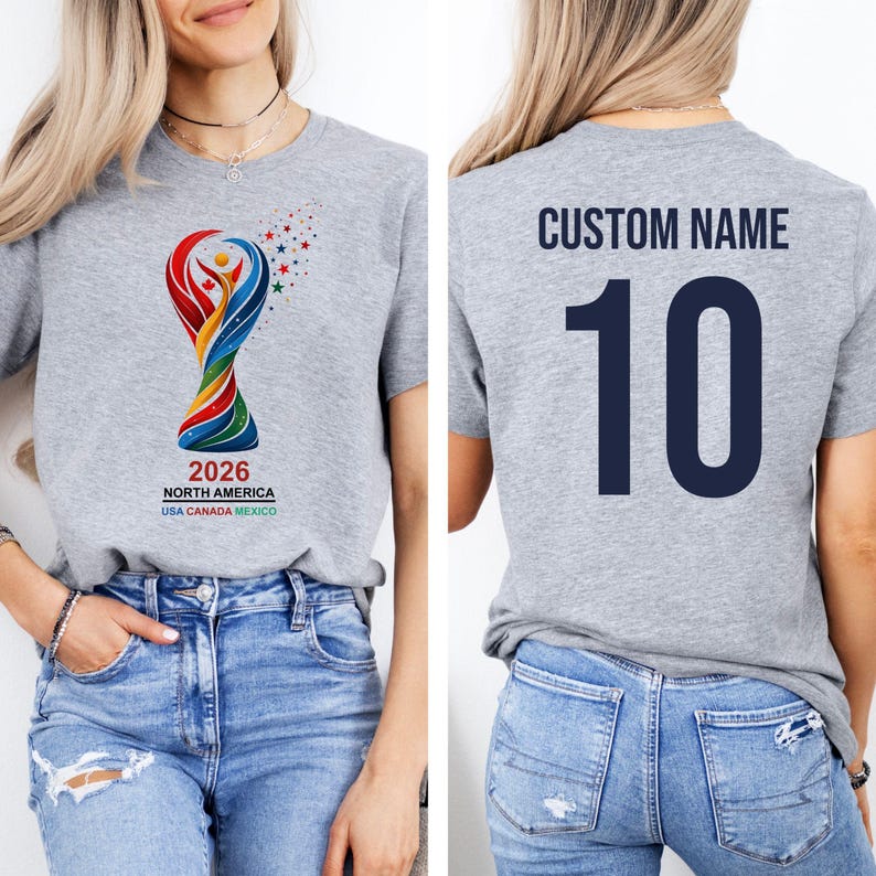 Op de afbeelding: Grijs t-shirt met een kleurrijke trofee-afbeelding en de tekst "2026 NORTH AMERICA USA CANADA MEXICO". De achterkant van het shirt toont "CUSTOM NAME" boven het nummer "10" in marineblauw. Korte mouwen.