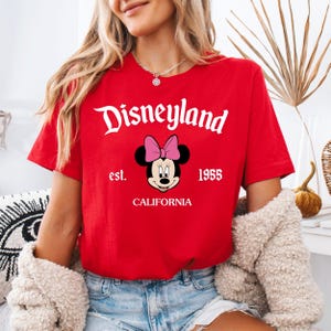 May include: A red t-shirt with the Disneyland logo in white script, a Minnie Mouse graphic, and the text "est. 1955 CALIFORNIA." The short-sleeved shirt is made of a soft material.