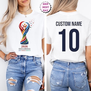 Op de afbeelding: Wit T-shirt met een kleurrijke afbeelding van een trofee en de tekst "2026 NORTH AMERICA USA CANADA MEXICO" op de voorkant. De achterkant van het shirt toont "CUSTOM NAME" boven het nummer "10" in marineblauw. Het model draagt een blauwe jeans.