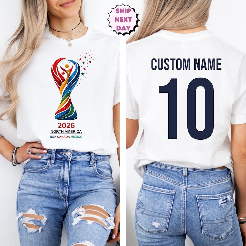 Op de afbeelding: Wit T-shirt met een kleurrijke afbeelding van een trofee en de tekst "2026 NORTH AMERICA USA CANADA MEXICO" op de voorkant. De achterkant van het shirt toont "CUSTOM NAME" boven het nummer "10" in marineblauw. Het model draagt een blauwe jeans.
