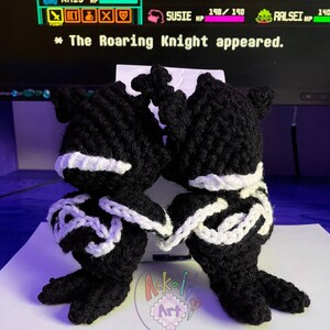 May include: Two handmade black and white crochet figures, possibly plush toys, are displayed. The figures have a distinctive design with white accents and are positioned close together. The background includes a computer screen with game-like text.