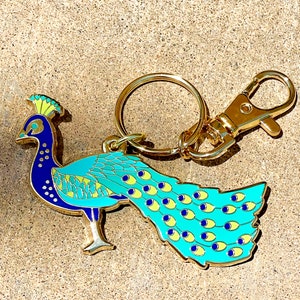 Puede incluir: Un llavero de metal dorado con forma de pavo real, con un mosquetón y un llavero. El pavo real tiene un cuerpo azul, plumas de cola turquesas con detalles amarillos y azules, y una cresta verde y amarilla. El llavero está sobre una superficie texturizada.