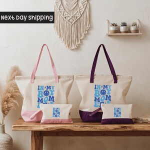 a set of two tote bags and a small bag on a wooden surface, with a wall decoration and some plants in the background.