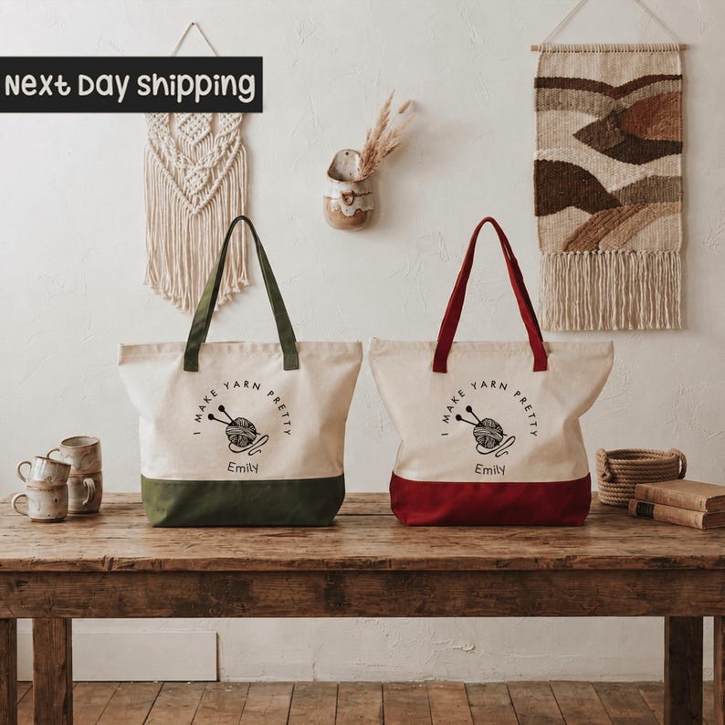 Two tote bags with the text "I make yarn pretty" and "I make yarn pretty Emily" printed on them, placed on a wooden table. The bags are green and red, and there are some decorative items like a woven wall hanging and a vase nearby.