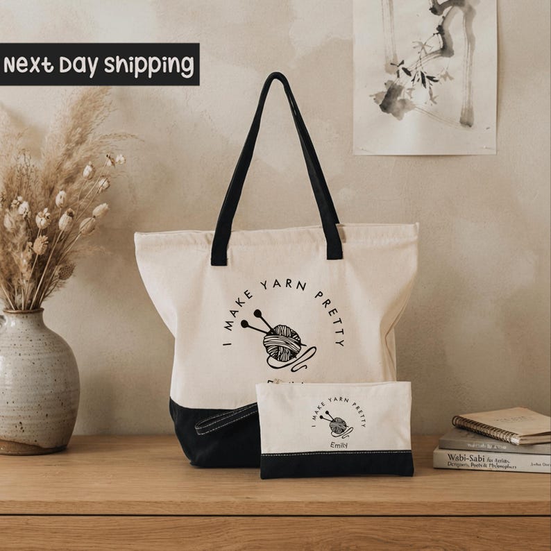 A beige tote bag with the text "I MAKE YARN PLETY" printed on it, along with a smaller bag and a vase of dried flowers on a wooden surface.