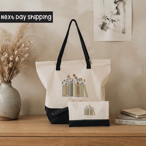 A white tote bag with a black handle and a smaller white bag with a black handle are displayed on a wooden surface, along with a vase of dried flowers and a framed picture.