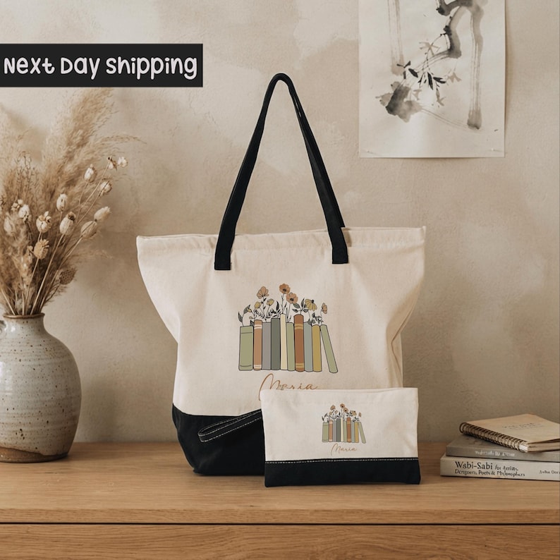 A white tote bag with a black handle and a smaller white bag with a black handle are displayed on a wooden surface, along with a vase of dried flowers and a framed picture.