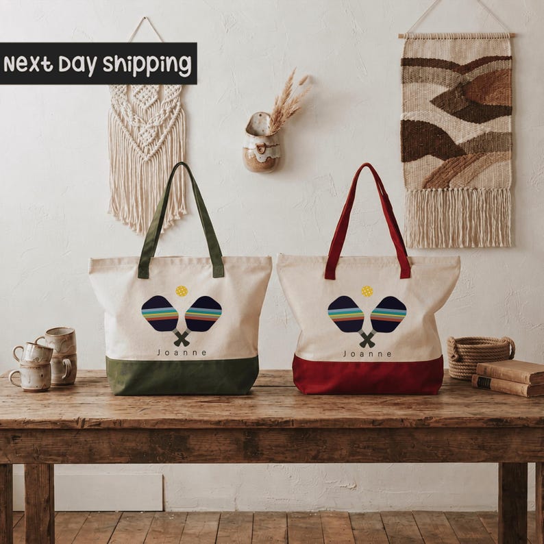 two tote bags with tennis rackets on them, placed on a wooden table. The bags are white and red, and they have the name "Joanne" printed on them. The table is set against a wall with a hanging tapestry, and there are some other items on the table, such as cups and a vase.