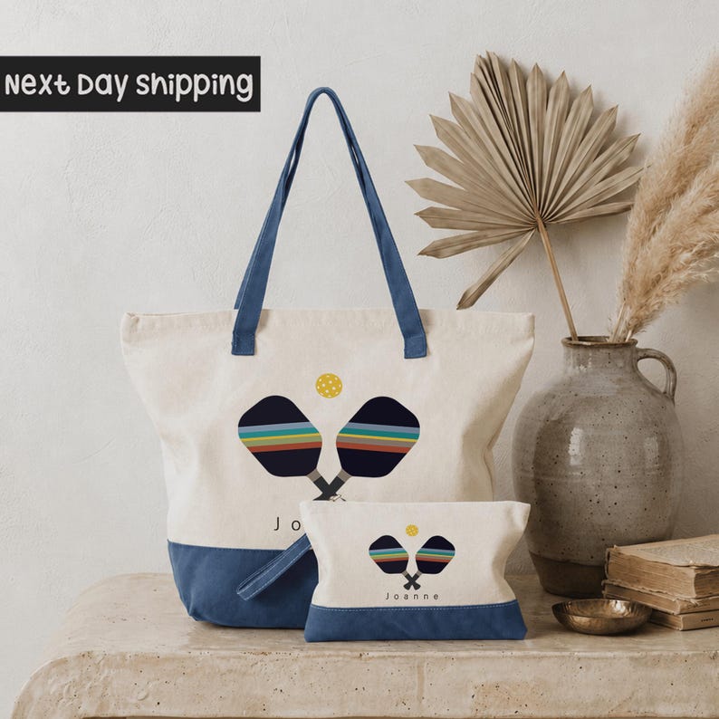 a white tote bag with a blue handle, a smaller bag with a similar design, and a decorative vase with a potted plant on a table.