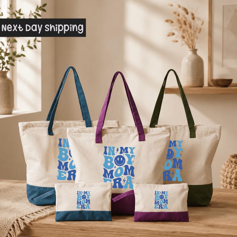 a collection of tote bags and small bags with the text "IN MY BOY MOM ERA" printed on them. The bags are arranged on a wooden surface, with a vase and some plants visible in the background.