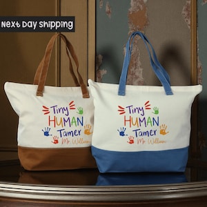 May include: Two canvas tote bags with the text "Tiny Human Tamer" in colorful letters. One bag has brown handles and a brown bottom, the other has blue handles and a blue bottom. Both bags have a white upper section.