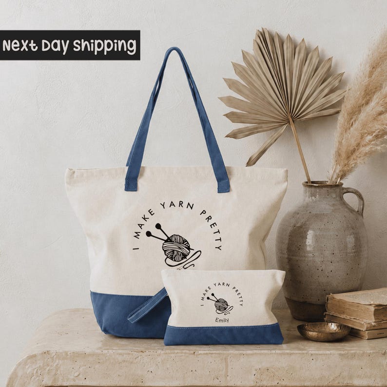 a large tote bag with a blue trim and a smaller bag with a blue trim, both with the text "I MAKE YARN PLETY" printed on them. The bags are placed on a table, and there is a vase with a potted plant and a stack of books nearby. The background of the image is a plain white wall.