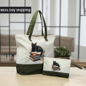 A white tote bag with a green trim and a smaller white bag with a cat design on it, both placed on a wooden surface.