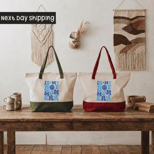 Two tote bags with text printed on them, placed on a wooden table against a wall with a woven wall hanging.