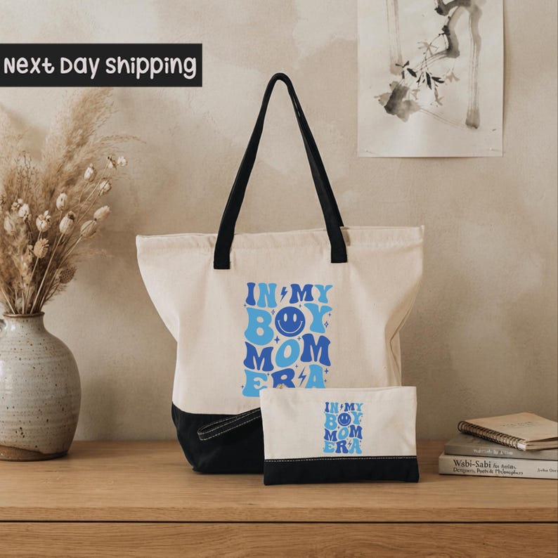 A white tote bag with a blue and black design, featuring the text "IN MY BOY MOM ERA" printed on it, placed on a wooden surface next to a vase of dried flowers and a stack of books.