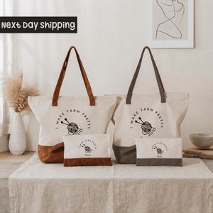 a set of two tote bags and a small bag, all with the text "I make yarn pretty" printed on them. The bags are placed on a table, and there is a vase and some books visible in the background.