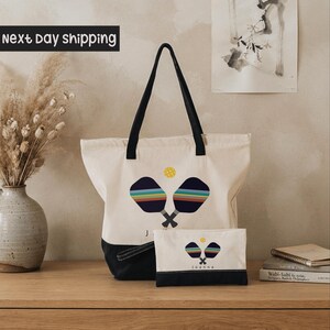 A white tote bag with a colorful tennis racket design and the name "Joanne" printed on it, placed on a wooden surface next to a vase of dried flowers and a stack of books.