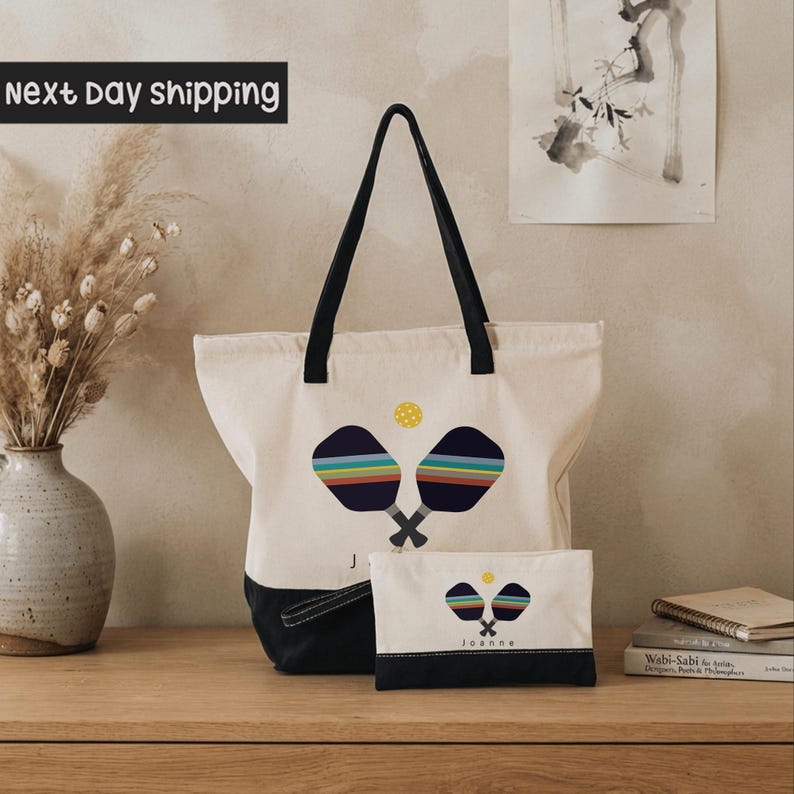 A white tote bag with a colorful tennis racket design and the name "Joanne" printed on it, placed on a wooden surface next to a vase of dried flowers and a stack of books.