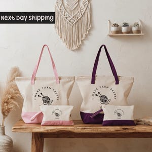 a set of two tote bags and a small bag, all with the text "I make yarn pretty" printed on them. The bags are white with purple handles, and the small bag is pink. The bags are placed on a wooden table, and there are some potted plants in the background.