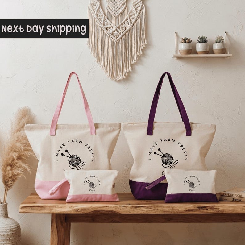 a set of two tote bags and a small bag, all with the text "I make yarn pretty" printed on them. The bags are white with purple handles, and the small bag is pink. The bags are placed on a wooden table, and there are some potted plants in the background.