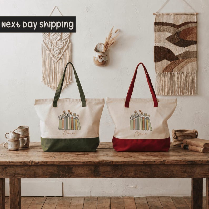 two tote bags with a floral design, placed on a wooden table. The bags are white with a red trim, and both have the word "Marina" printed on them. The table also has a few items on it, including a vase, a cup, and a basket. The background includes a wall with a hanging tapestry and a shelf with various items.