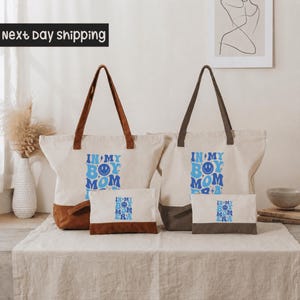 two tote bags with the text "IN MY BOY MOMM" printed on them, along with a small bag and a picture frame on a table.