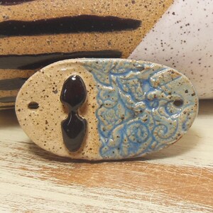 May include: A ceramic oval-shaped tile with a blue and white floral design on one side and a tan speckled glaze on the other side. A black glaze is applied to the centre of the tan side in the shape of a teardrop.