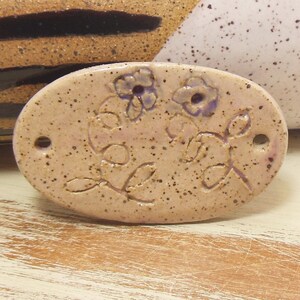 May include: A light brown oval ceramic tile with a raised design of two purple flowers and leaves. The tile has a speckled texture.