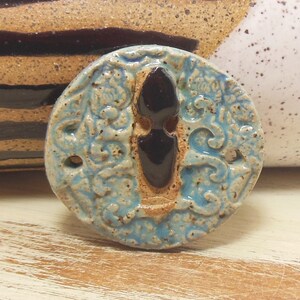 May include: A round ceramic button with a blue and brown glaze. The button features a raised design of a stylised figure with a keyhole shape. The button has two holes for sewing.